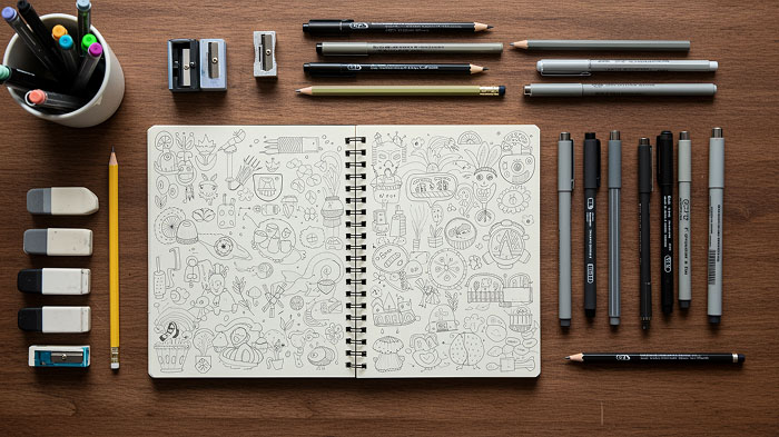 A neatly arranged flat lay of essential doodling tools on a wooden desk, featuring a sketch pad with in-progress doodles, pens, pencils, markers, erasers, and blending stumps.