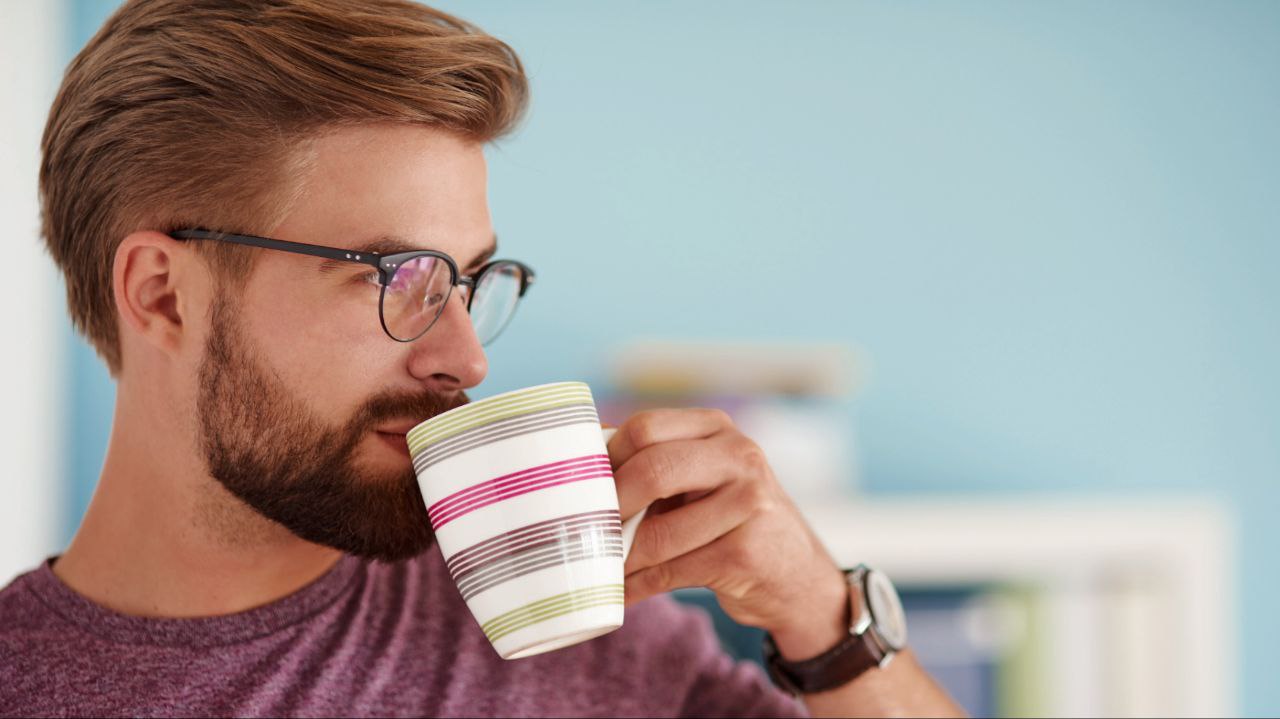 A man wearing glasses, a t-shirt, and a watch, drinking from a colorful mug